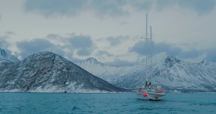 A sailboat is sailing towards a mountainous coastline on a winter day