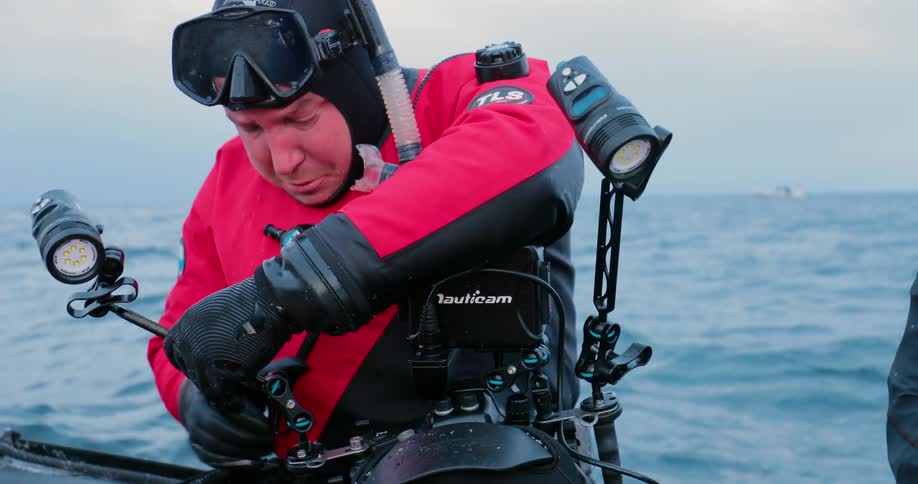 A man in diving gear is adjusting the lights on his camera as he sits in a dinghy