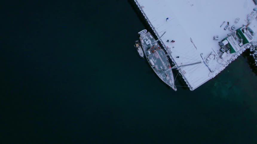 People walk and stand on a snow-covered dock near a sailboat during the day