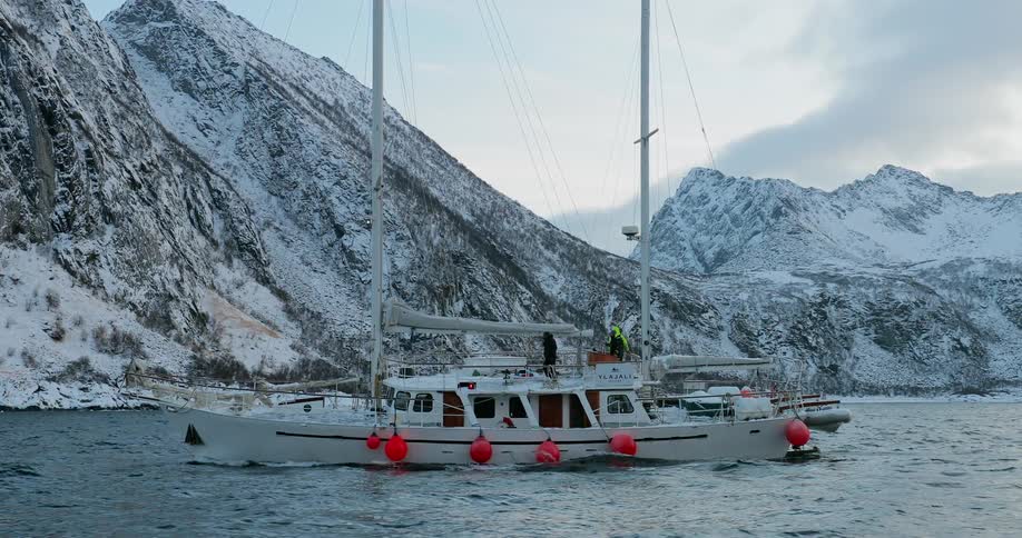 A sailboat is sailing off a mountainous coastline on a winter day