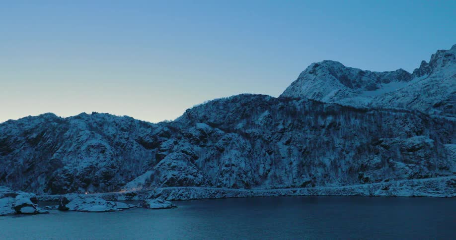 A vehicle is driving on a coastal road past snow-covered mountains at dawn