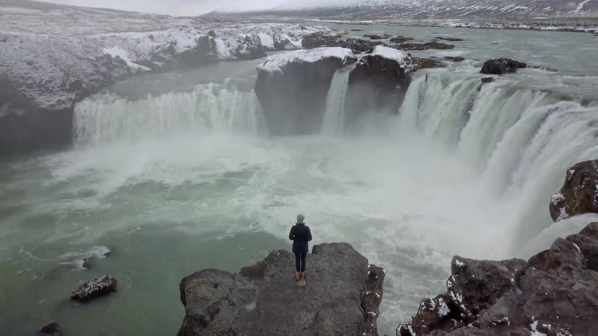 A woman stands atop a rock overlooking the rapidly flowing water