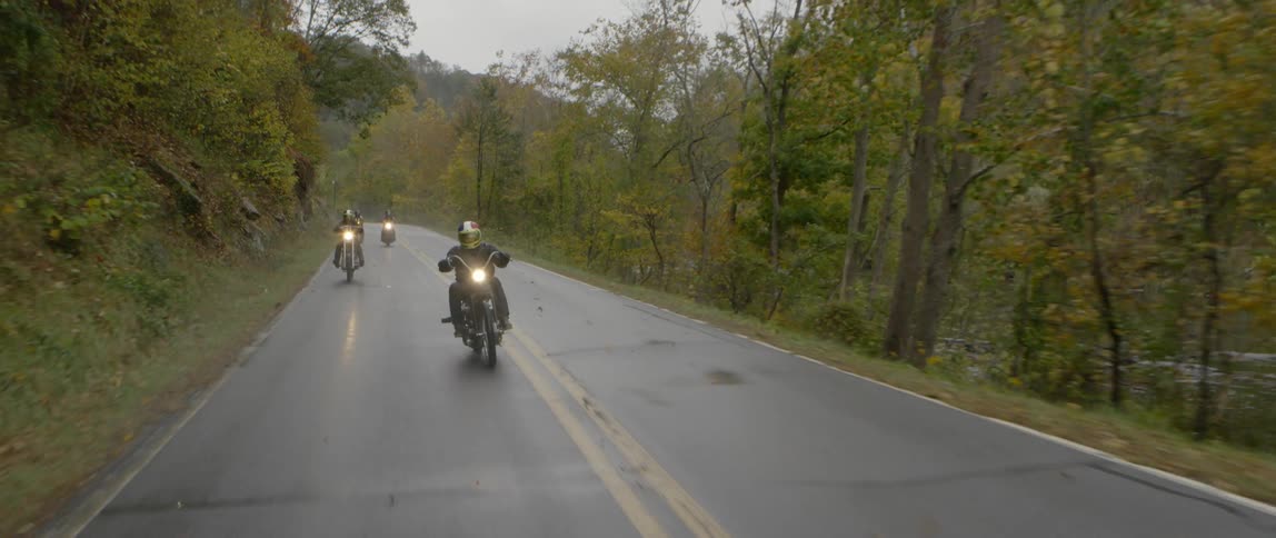 Four people ride motorcycles down a curved wet road lined with trees