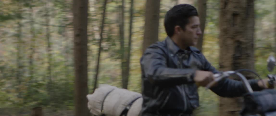A man is riding a motorcycle into a campsite in a forest during the day