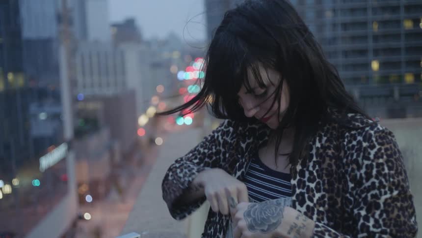 Woman stands on a rooftop in a city and puts a piece of paper in her pocket