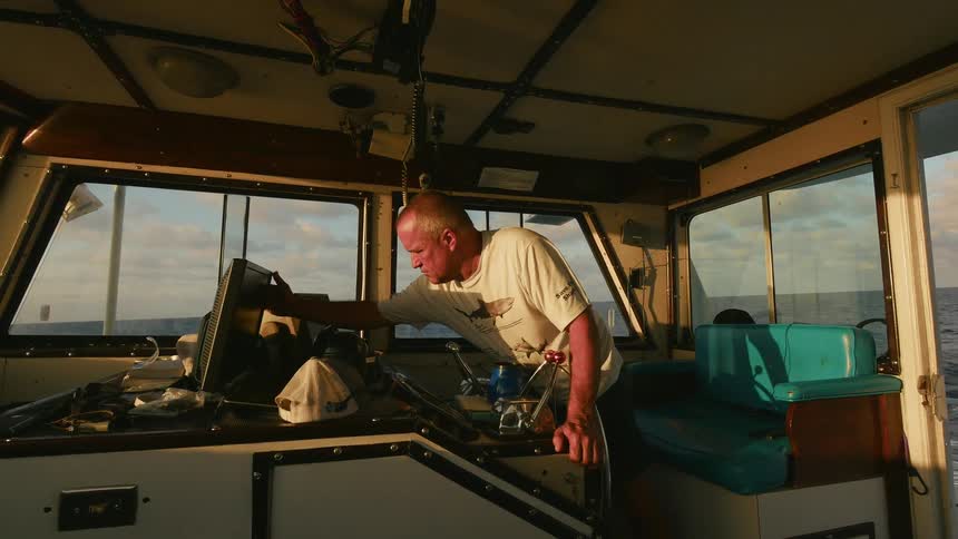 A man stands at the controls of a ship as it sails on a cloudy day