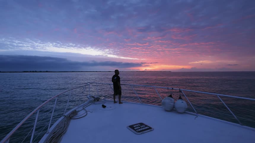 A man stands on the deck of a large ship as it sails on the ocean during sunset