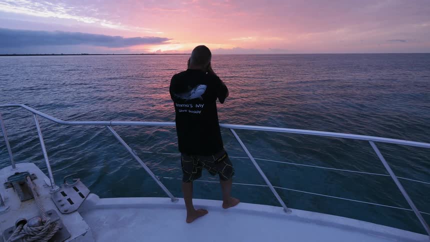 A man is standing at a railing on a boat taking a photo at sunset