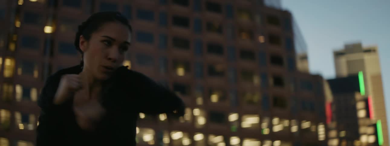 A woman shadow boxes on a city rooftop at dusk