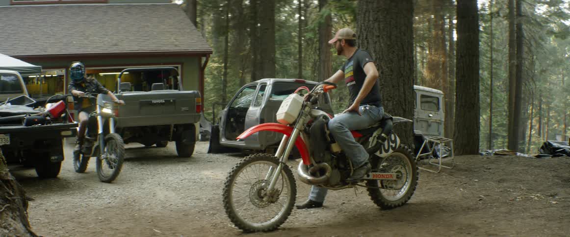 A man sitting on a motorcycle watches a woman on a motorcycle pulls past him