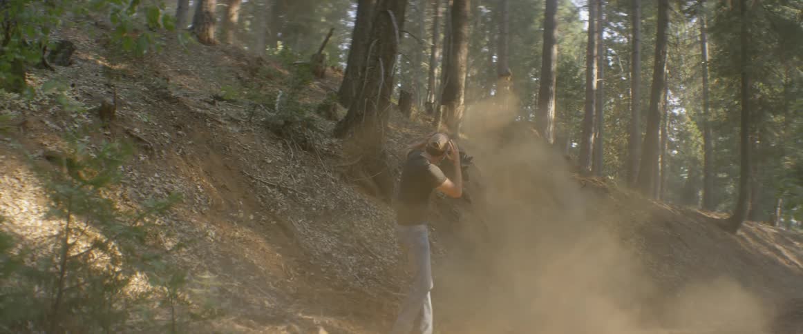 A man watches a woman ride a bike up a ramp in the woods