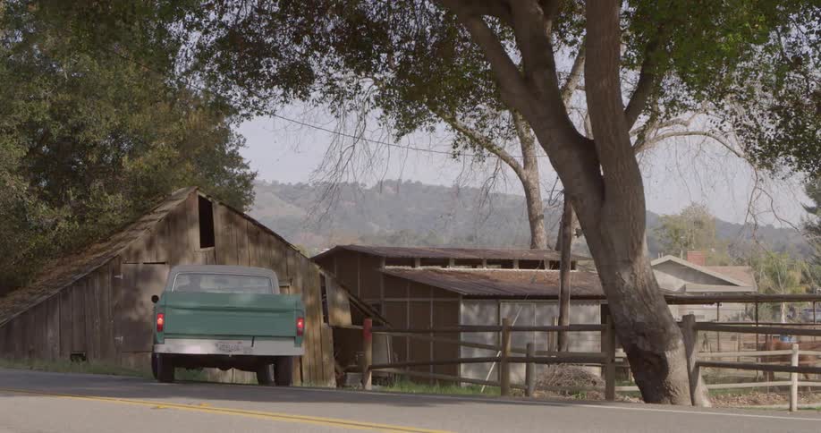 A blue truck drives to a barn under a tree