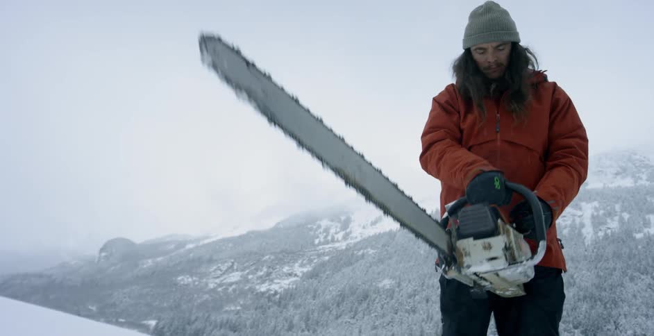 A man standing in a snow-covered natural setting is trying to start a chainsaw