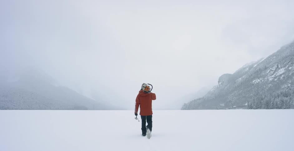 A man carries a fishing pole and chainsaw through the snow