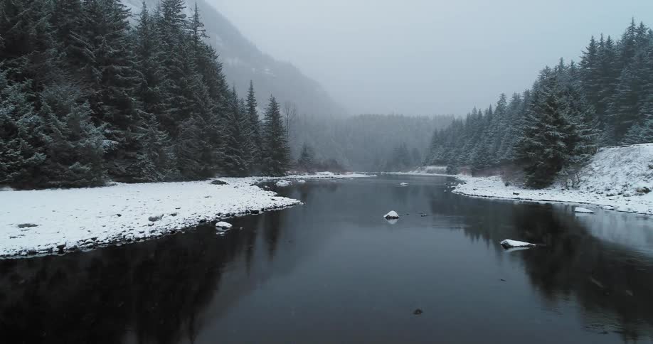 Snow flurries are falling over a river running through a natural setting