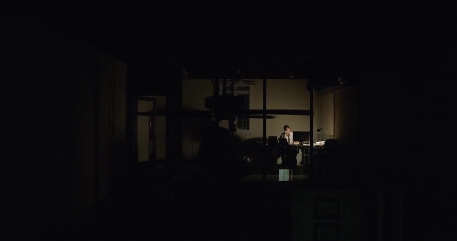 A man sitting at his desk with the lamp on can be seen through glass doors
