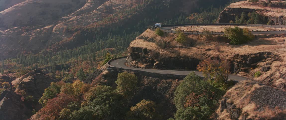 A truck is parked at the top of a ridge overlooking the road and forest below