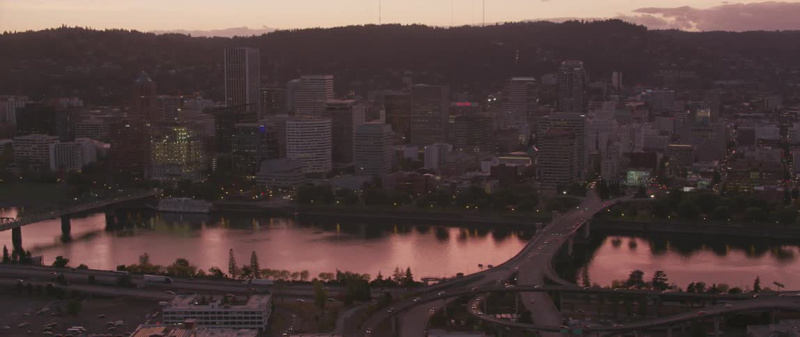 Traffic is flowing on a highway complex across a river from a city at dusk
