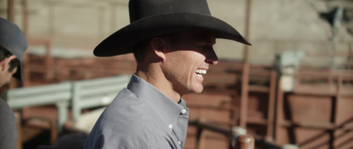 A cowboy is smiling as he stands outside at a rodeo during the day