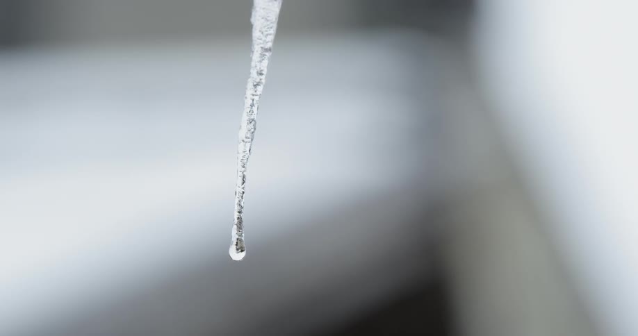 A drop of water slowly falls from a small icicle in the rain