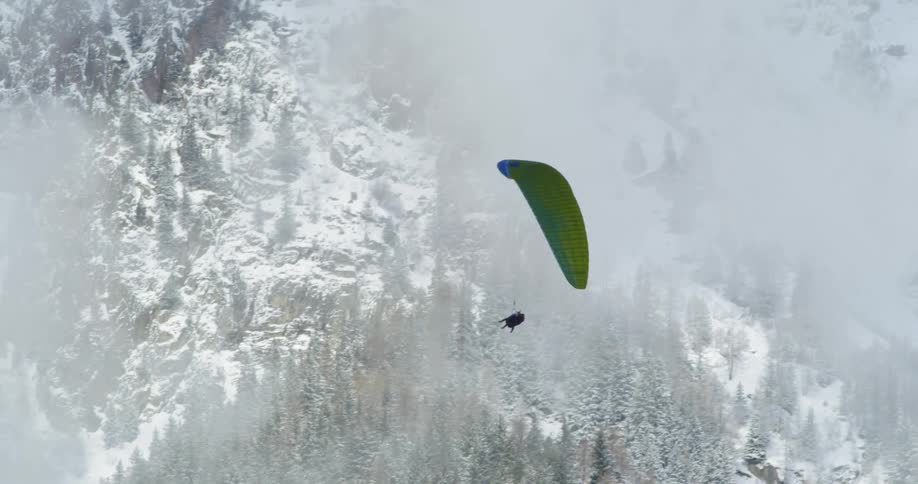 A person paraglides over trees and snow covered slopes
