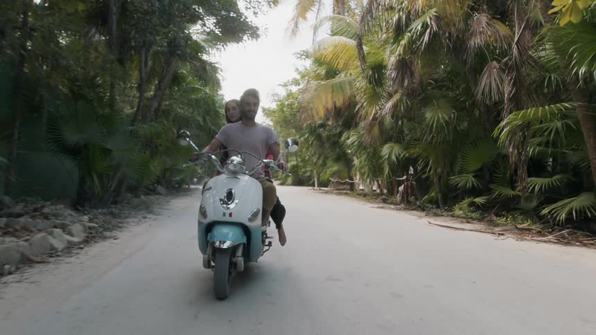 A couple are riding a moped on a road in a tropical setting during the day