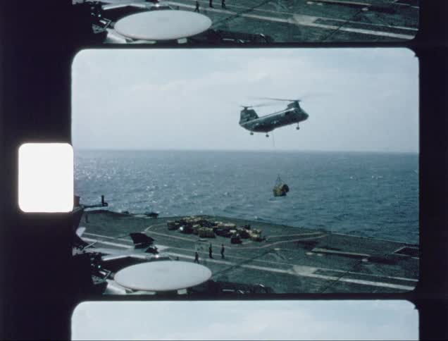 Military helicopter lowering cargo onto an aircraft carrier's deck on a cloudy day