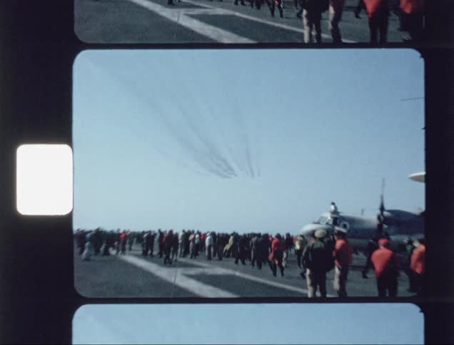 People on an aircraft carrier watch jets fly overhead