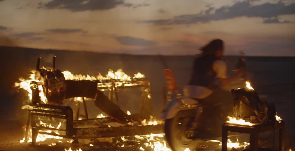 A person races a motorcycle through burning debris