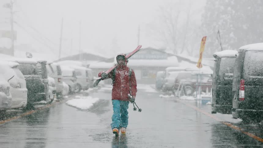 A skier carries skis in a parking lot as it snows