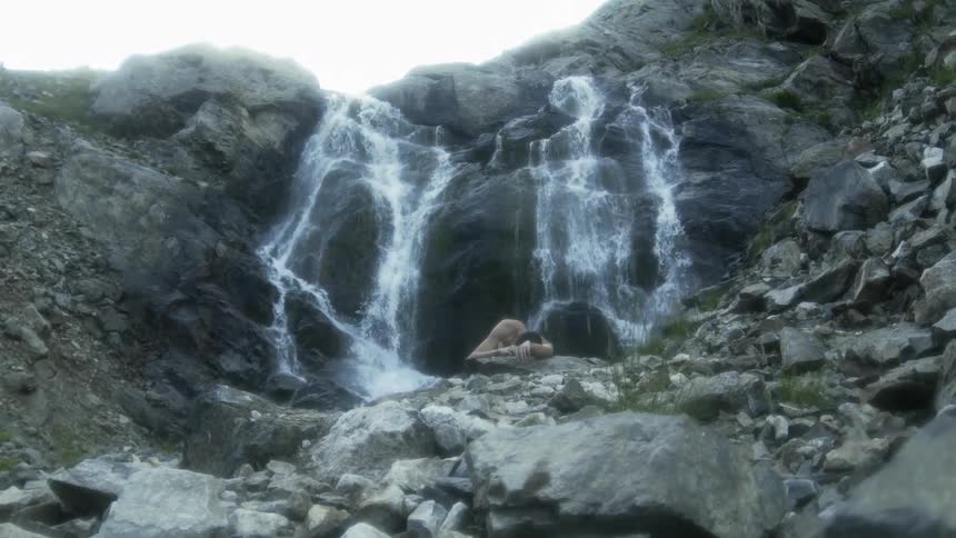 A naked man is lying on rocks near a waterfall in daylight