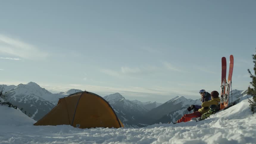 Two skiers are resting at a campsite on a hillside during the day