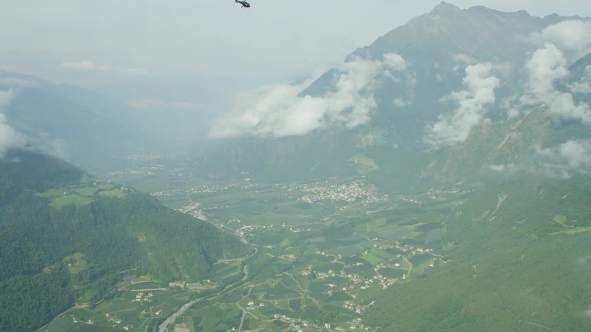 A helicopter flies above a valley town between tall mountains