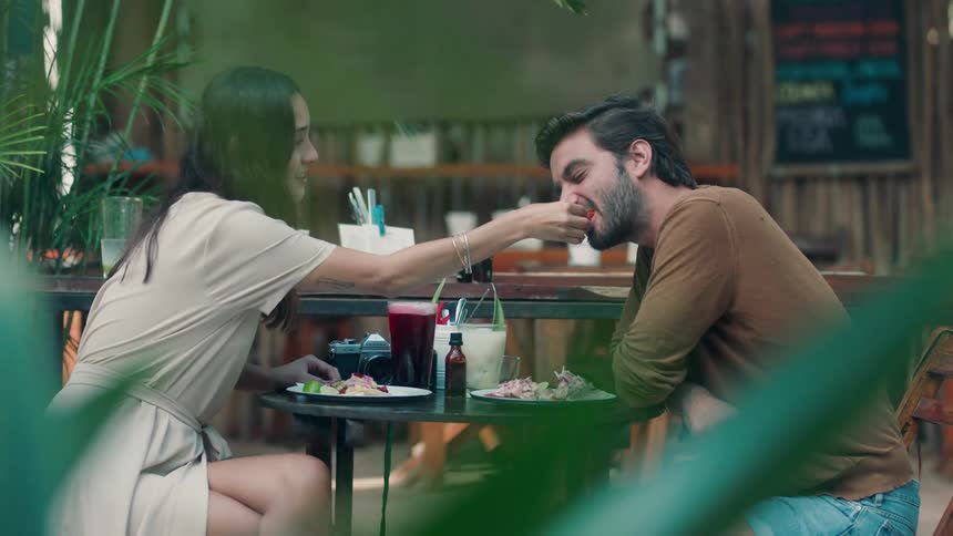 A woman is feeding a man a bite of her food as they sit at an outdoor restaurant