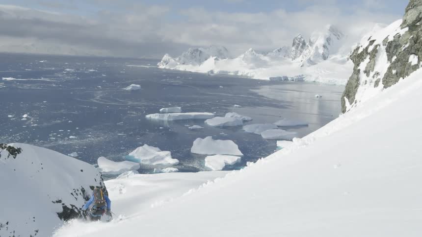 Man wearing a backpack skis down a snowy mountain towards a lake