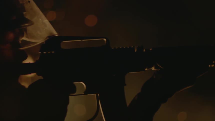 A person aims a gun while a fire burns behind them