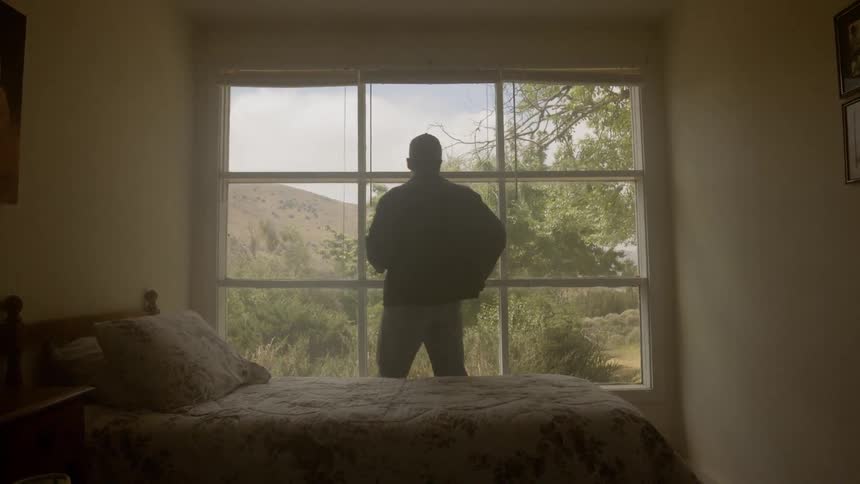 A man walks around the bed and looks out the large window