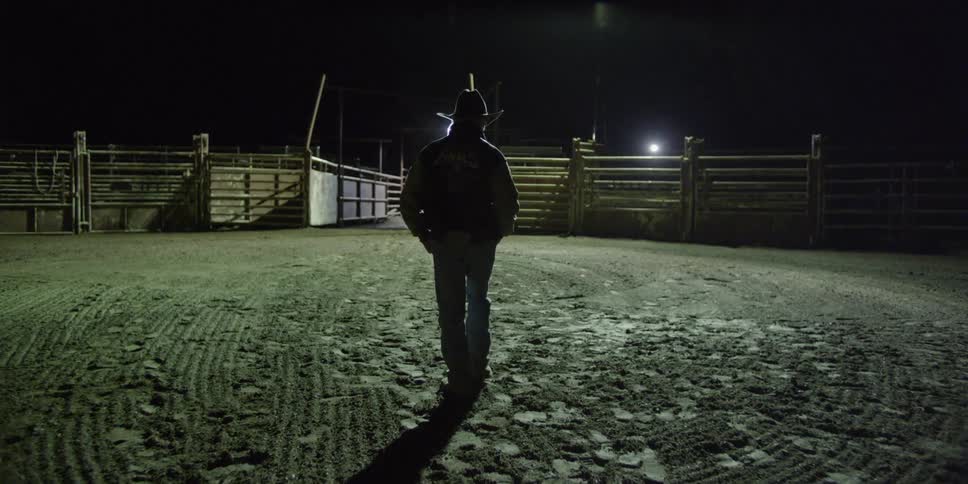 A cowboy walks through a paddock at night and stops in the center