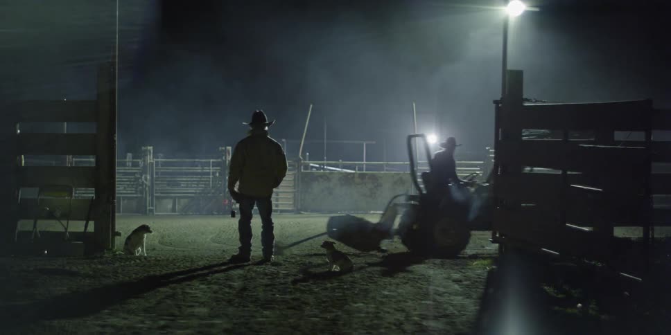 A man is standing in the gate of a rodeo arena watching preparations at night