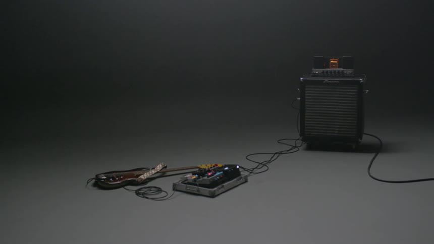 A bass guitar with a pedal board and amplifier sit in a dim room