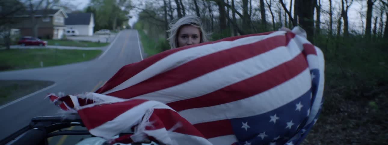 A woman holds an american flag out of a driving vehicle