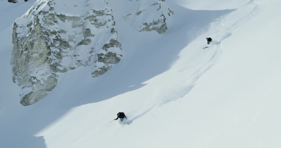 Two skiers are skiing down a fresh mountain slope during the day