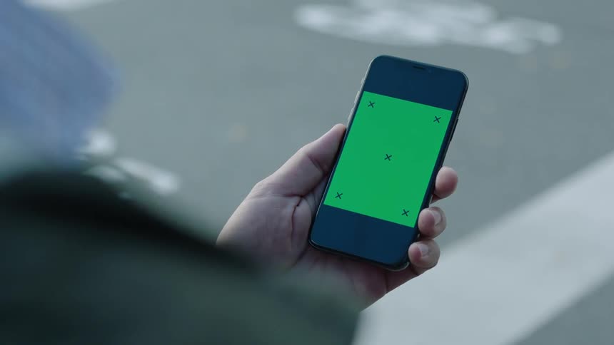 Cinematic close-up of a person standing by a street holding a smartphone with a green screen and tracking markers in daylight. The phone stays still without interaction, perfect for VFX, screen replacement, app mockups, and compositing.