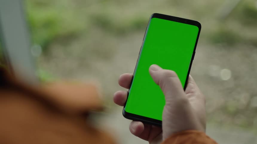 Close-up of a person holding a smartphone with a green screen outdoors, swiping across the display with their thumb multiple times. Filmed in natural light, ideal for VFX, screen replacement, app mockups, and digital interaction projects.