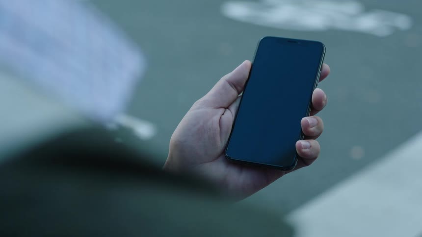 Close-up of a person standing by the roadside holding a smartphone with a black screen. Filmed outdoors in natural daylight, ideal for VFX, screen replacement, app mockups, digital lifestyle, and technology-related projects.