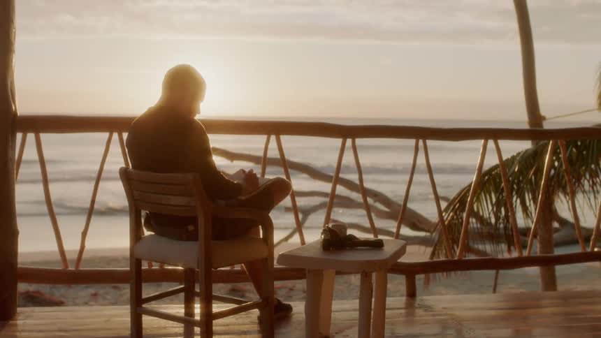 A man writes in a notebook on a porch on the beach