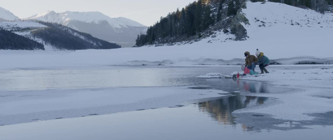 A family is pushing toy boats into the stream of a river on a winter day