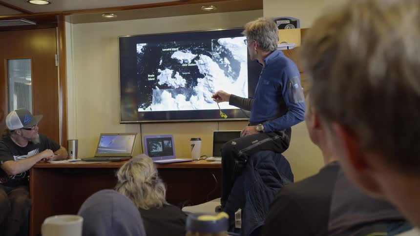 A man instructs other people using a pointer and a television screen