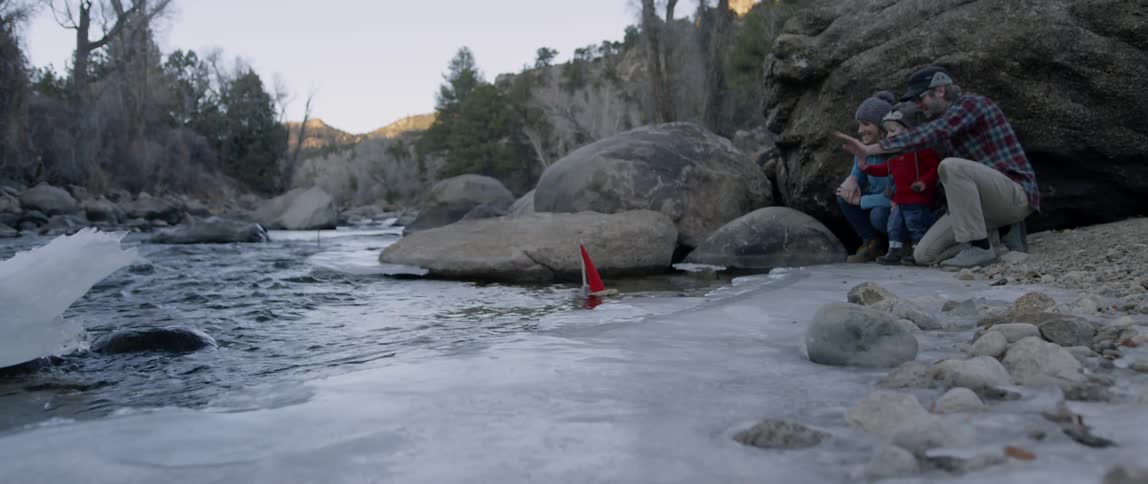 Couple and child waving at a toy boat that they launched in an icy river