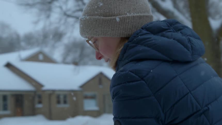 A woman is shoveling snow outside in a suburban setting as it snows